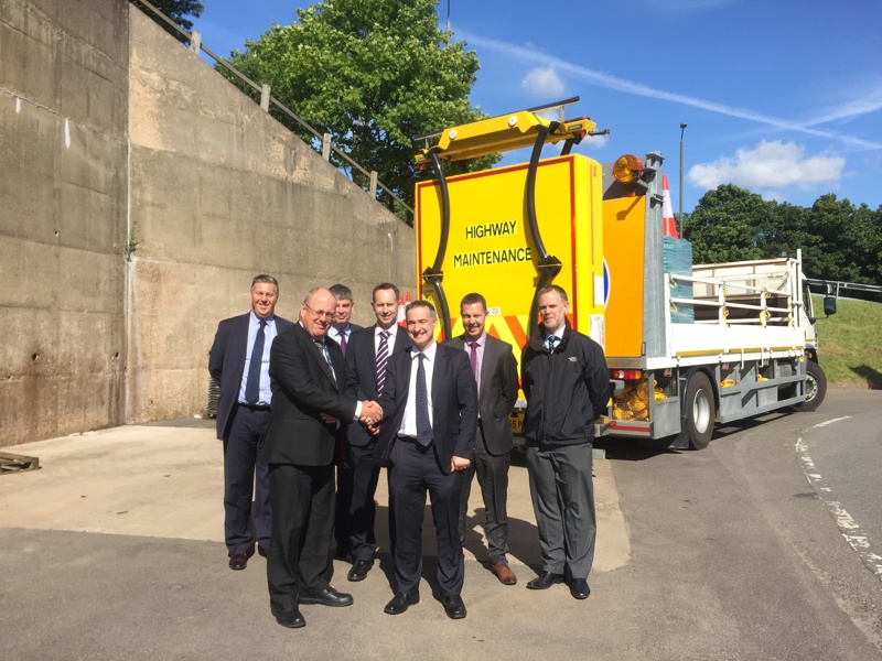 Group of men in formal attire, stood in front of a highways vehicle. 