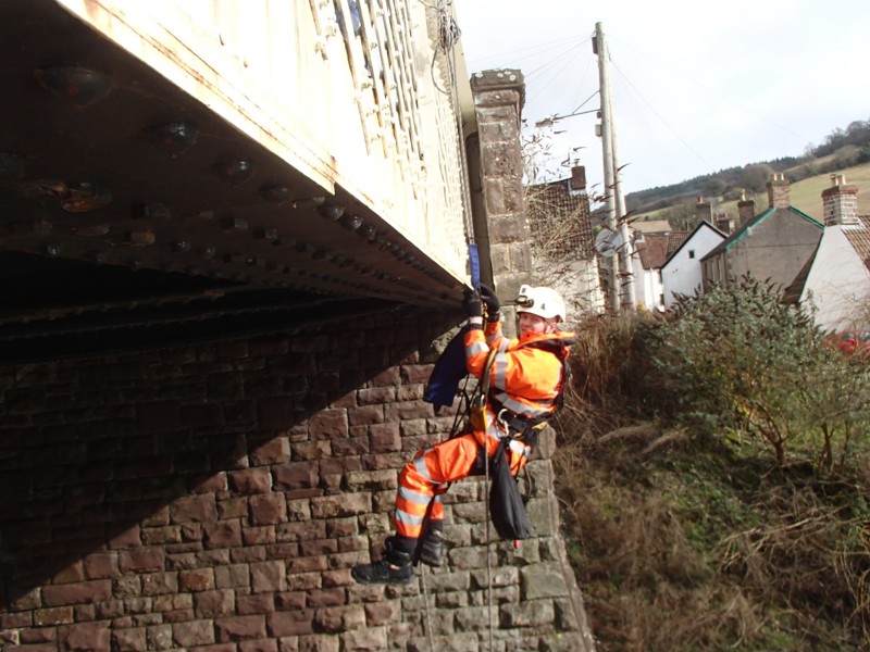 Image of a man wearing PPE and a harness hanging from a bridge.