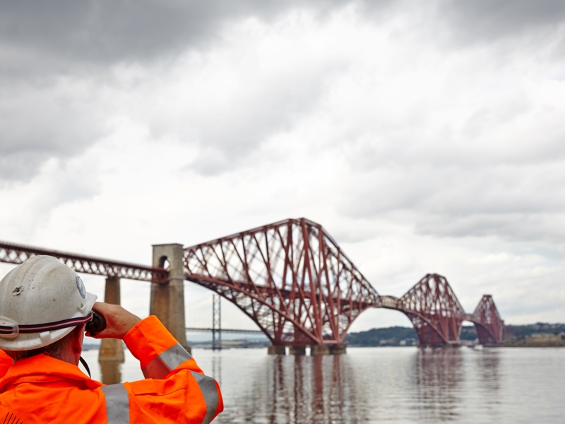 A man in PPE, looking through binoculars at a bridge. 