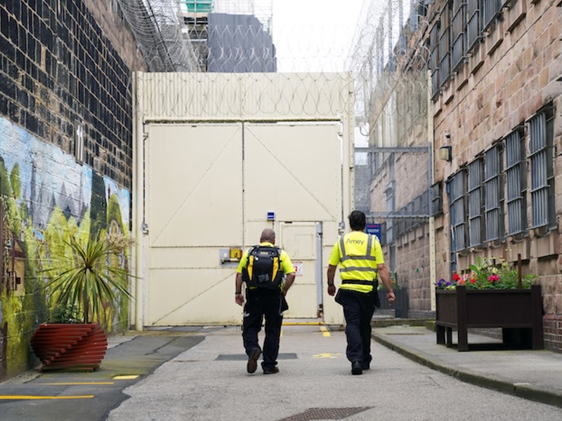 Two Amey employees in high-visibility clothing walk towards a secure gate within a prison complex, with barbed wire overhead and colourful murals and flower planters brightening the surroundings.