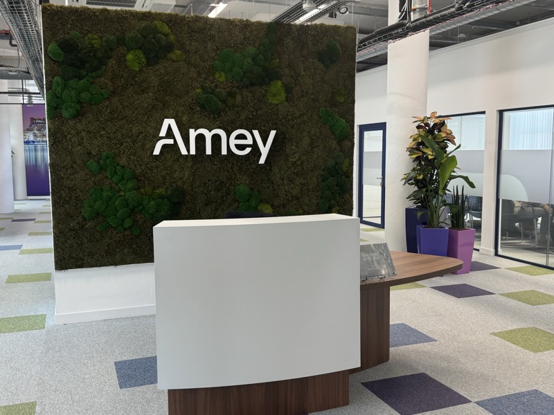 A picture of the newly refurbished Amey reception desk at Liverpool’s Matchworks, featuring a moss-covered wall with the Amey logo, a modern curved desk, and colourful square-patterned carpet tiles.