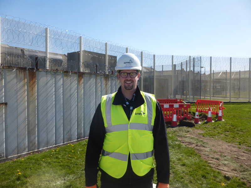 Image of an Amey employee in PPE, stood outside of a secured fence.