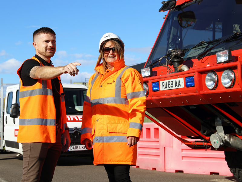 Image of a male and female in PPE, stood in front of vehicles. 