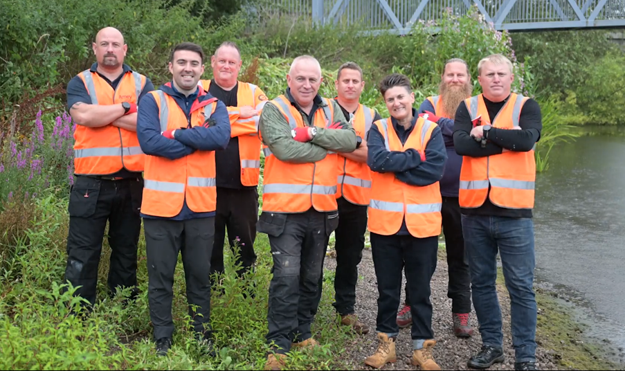 A group of men and women standing beside a river wearing orange PPE.