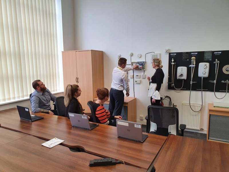 Image of people sat round a desk, watching two people inspect demo showers on a wall.