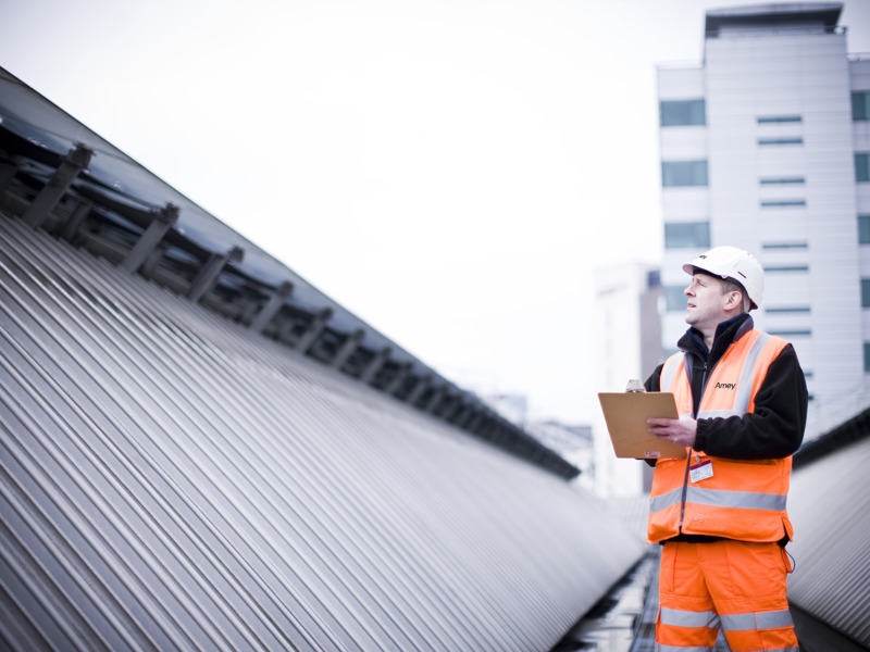 A male engineer in high-visibility Amey workwear and a hard hat inspects rooftop infrastructure with a clipboard, in an urban setting.
