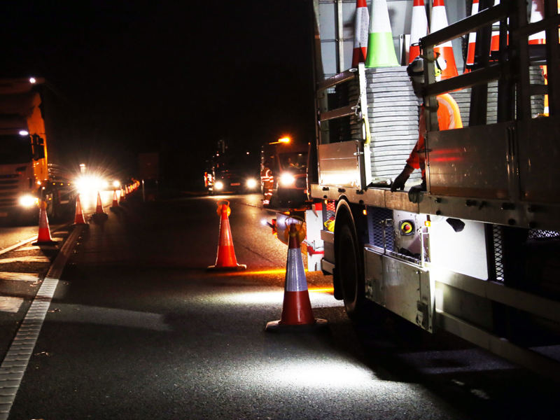 Night-time roadworks on a motorway with a maintenance truck, traffic cones, and Amey workers in high-visibility clothing.