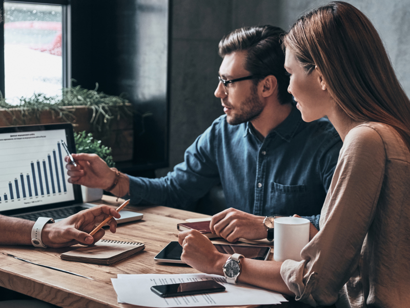 Two men and a woman sharing a laptop screen at a wooden table.