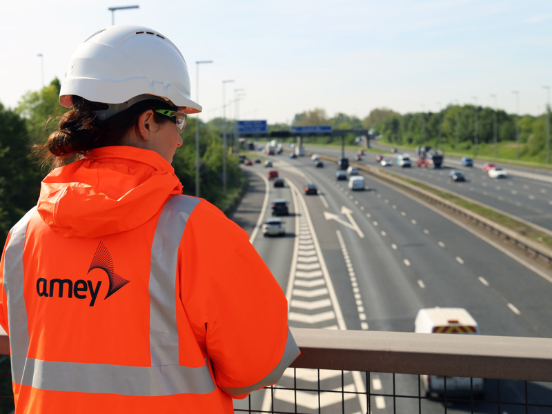 Image of a female Amey employee wearing PPE, standing with their back o the camera on a bridge.
