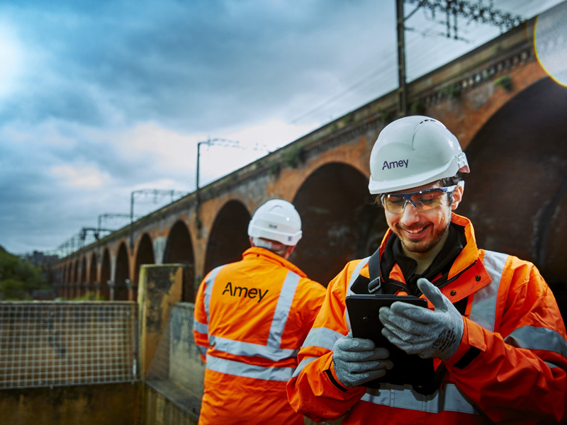 Two Amey rail workers in high-vis gear inspect infrastructure near a brick viaduct, with one smiling while using a tablet.