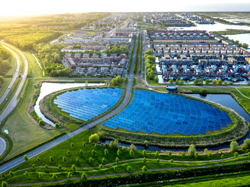 Aerial view of a solar farm with rows of blue solar panels surrounded by green grass and water features, located near a modern residential neighborhood with neatly aligned houses and roads. The area is bathed in soft sunlight, creating a vibrant and sustainable landscape.