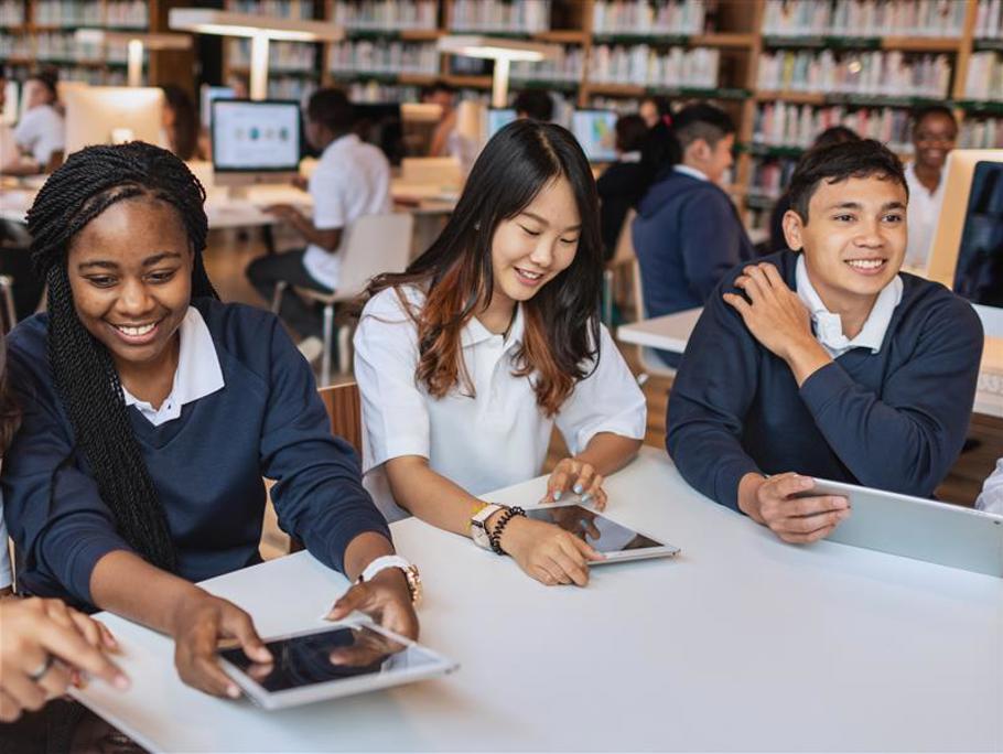 Three students seated at a table. Two of them are using electronic tablets.