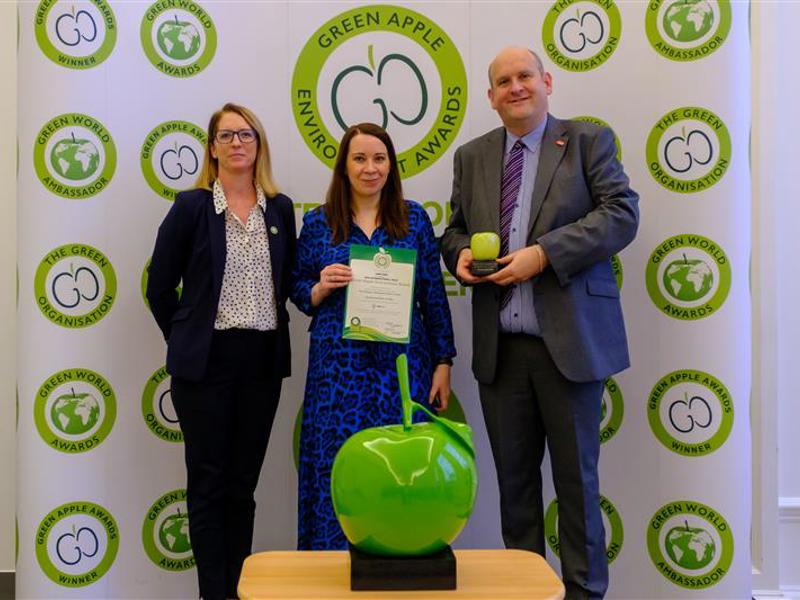 Three people standing in front of a Green Apple Environment Awards backdrop, holding a certificate and a trophy. In the foreground, a large green apple trophy is displayed on a table. The individuals are dressed formally, smiling at the camera.