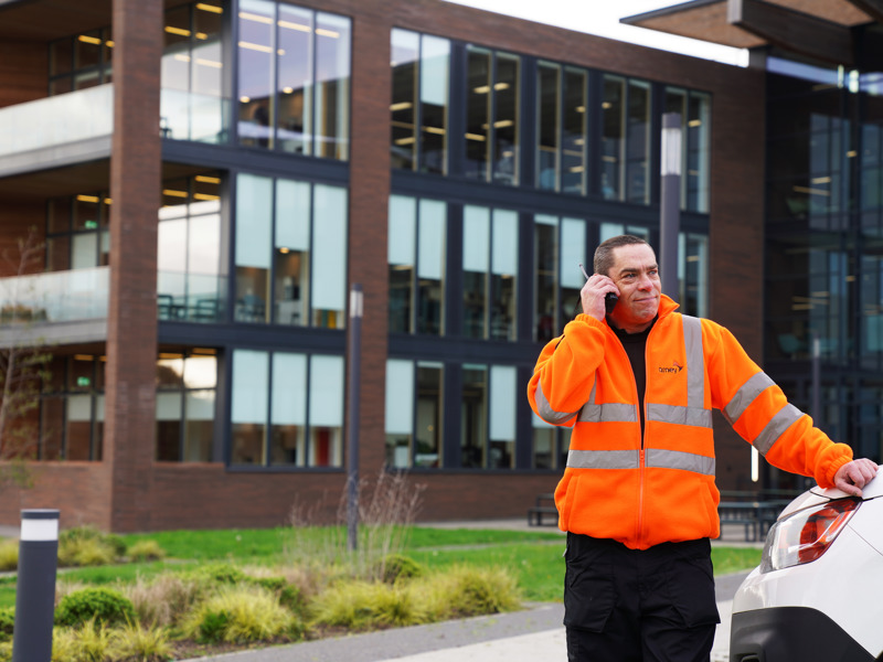 A male in an orange PPE jacket, on a radio stood in front of a glass building.